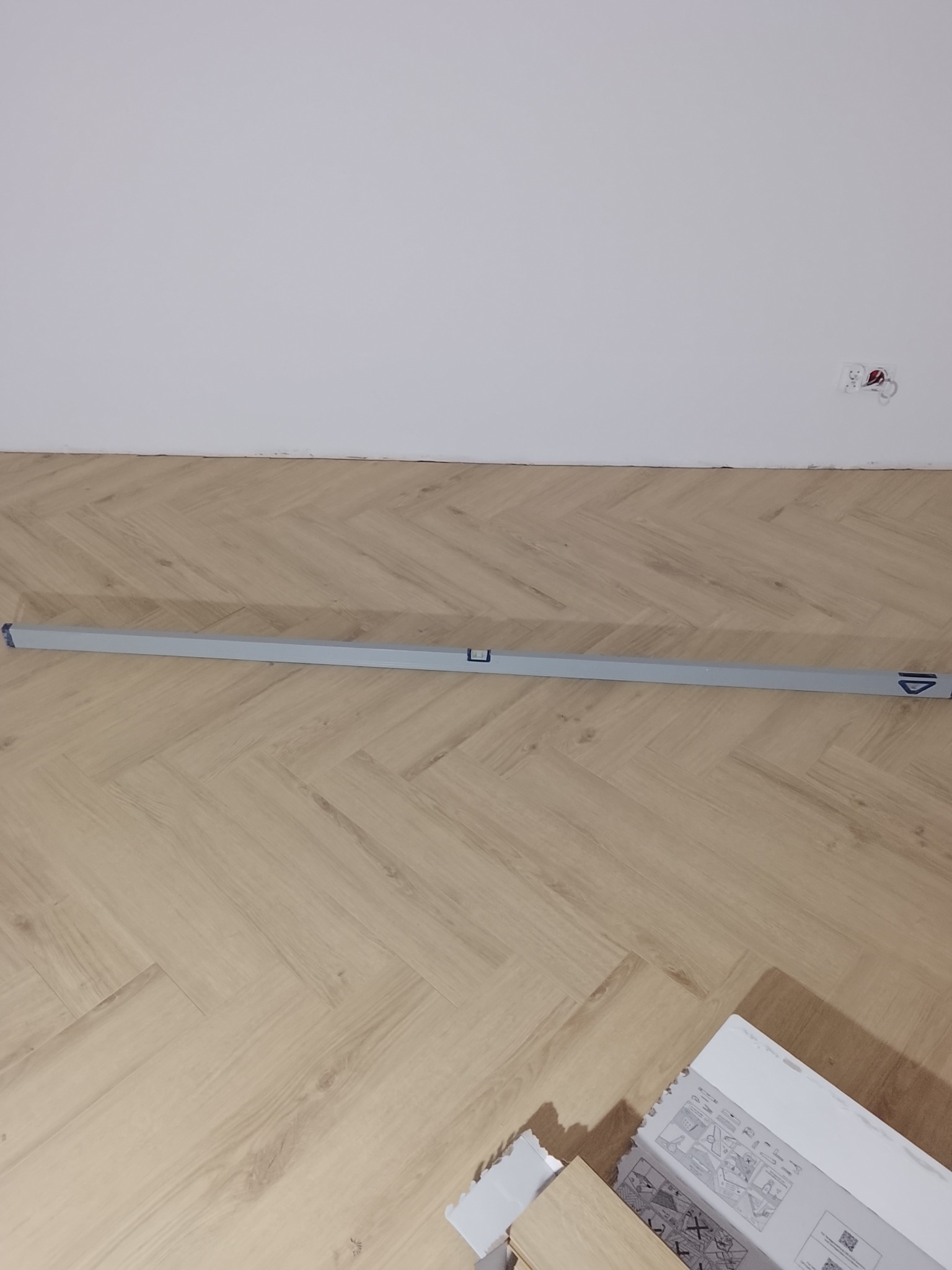 Ułożona podłoga w jodełkę w Nowym Kozłowie Drugim, sprawdzana poziomicą. Widoczne opakowania paneli. Biała ściana z gniazdkiem elektrycznym.