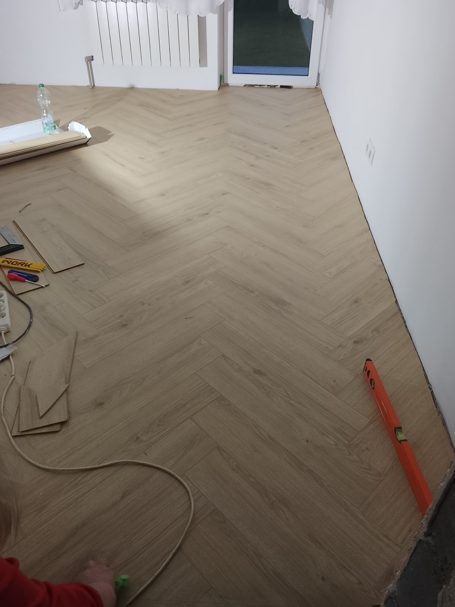 Ułożona podłoga z paneli w jodełkę, widoczny poziomica przy ścianie, narzędzia i resztki paneli. Jasne wnętrze z oknem w tle.