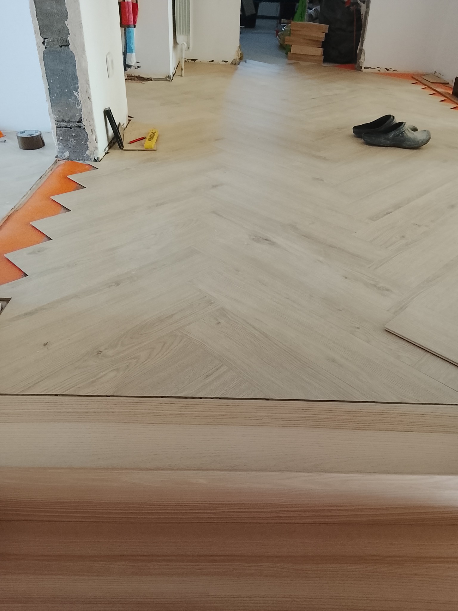 Układanie jasnych paneli podłogowych w jodełkę, widoczne narzędzia i fragment podkładu. Etap prac wykończeniowych w jasnym wnętrzu.