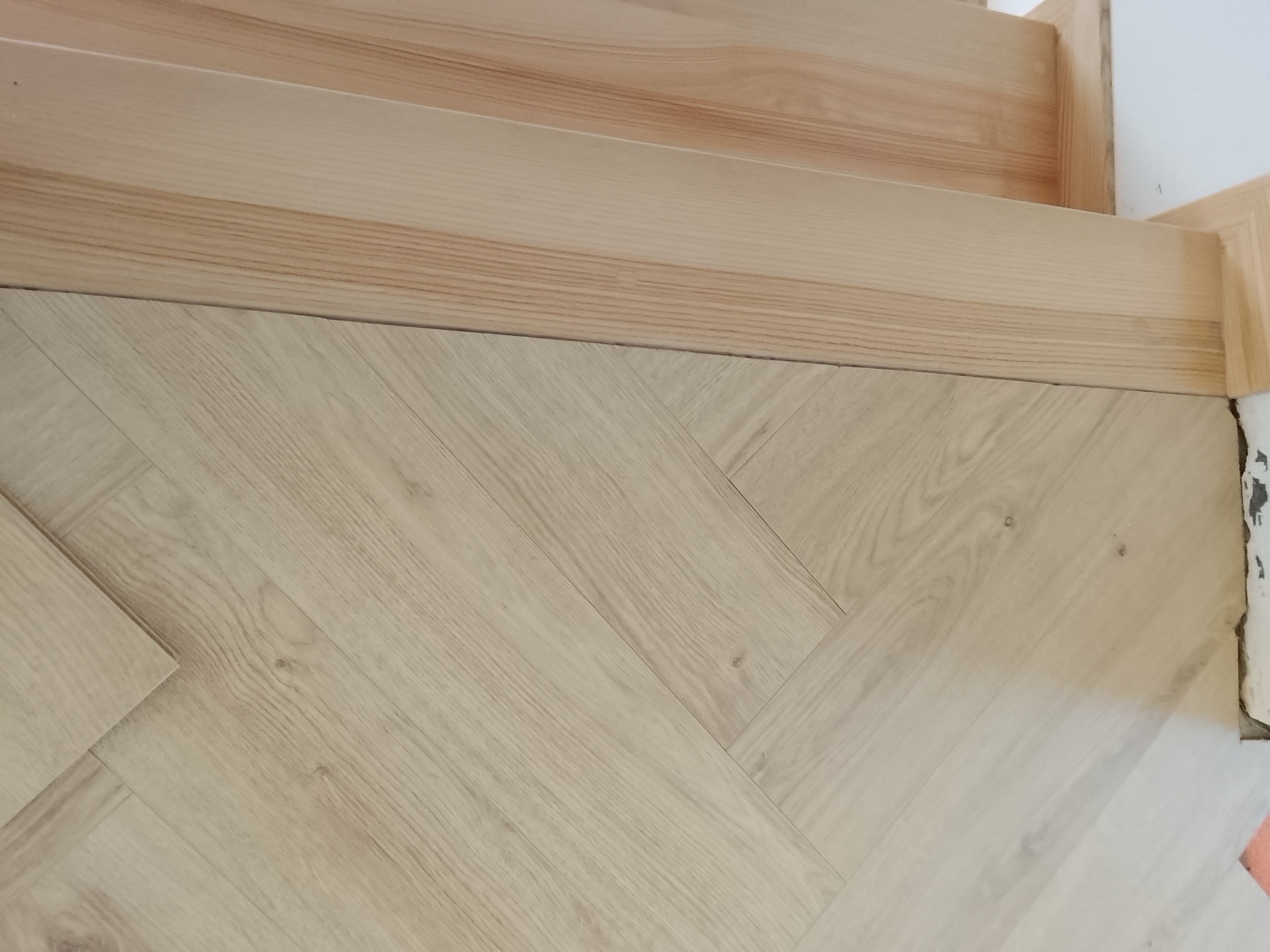 Jasna podłoga ułożona w jodełkę, z widocznymi drewnianymi listwami przypodłogowymi i fragmentem schodów. Ujęcie z bliska, skupione na detalu wykończenia.