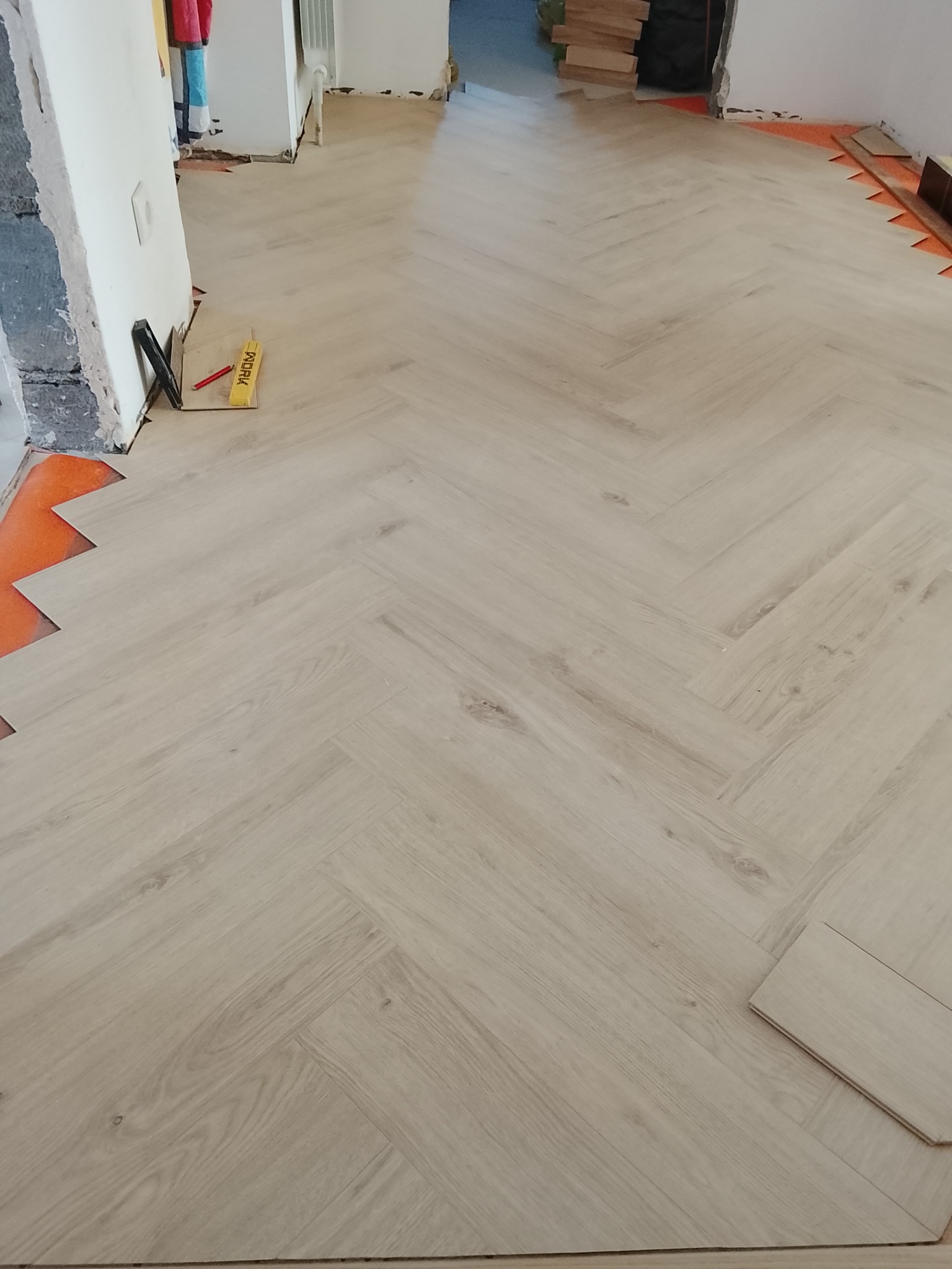 Jasna podłoga ułożona w jodełkę, widoczne narzędzia glazurnicze i fragmenty ścian w trakcie wykończenia. Na podłodze leżą panele.