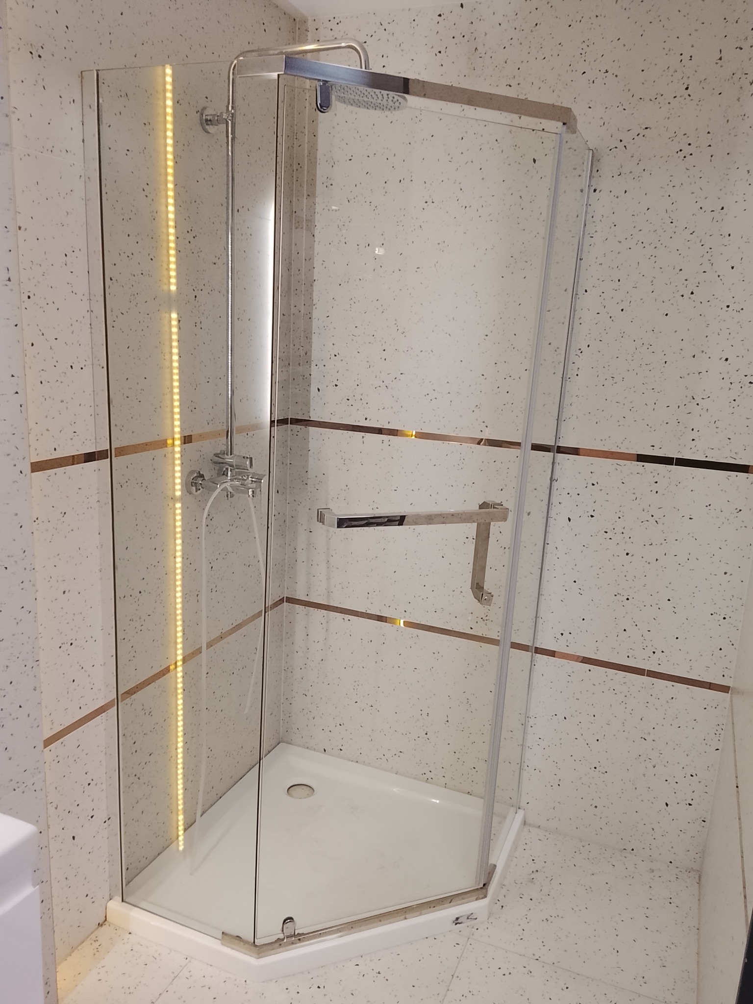 Szklana kabina prysznicowa w narożniku łazienki z mozaikową ścianą i złotymi akcentami. Widoczny chromowany zestaw natryskowy i uchwyt. Nowoczesny design.