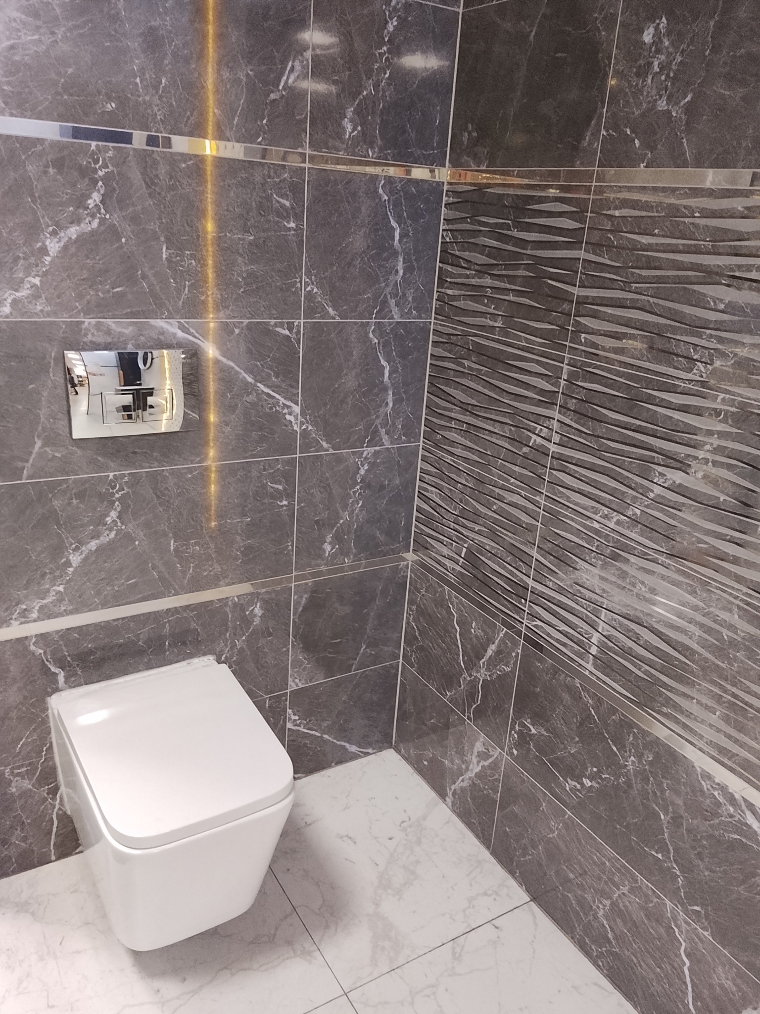 Nowoczesna łazienka z szarymi płytkami imitującymi marmur, dekoracyjną ścianą z falistym wzorem i podwieszaną toaletą. Chromowane listwy wykończeniowe.