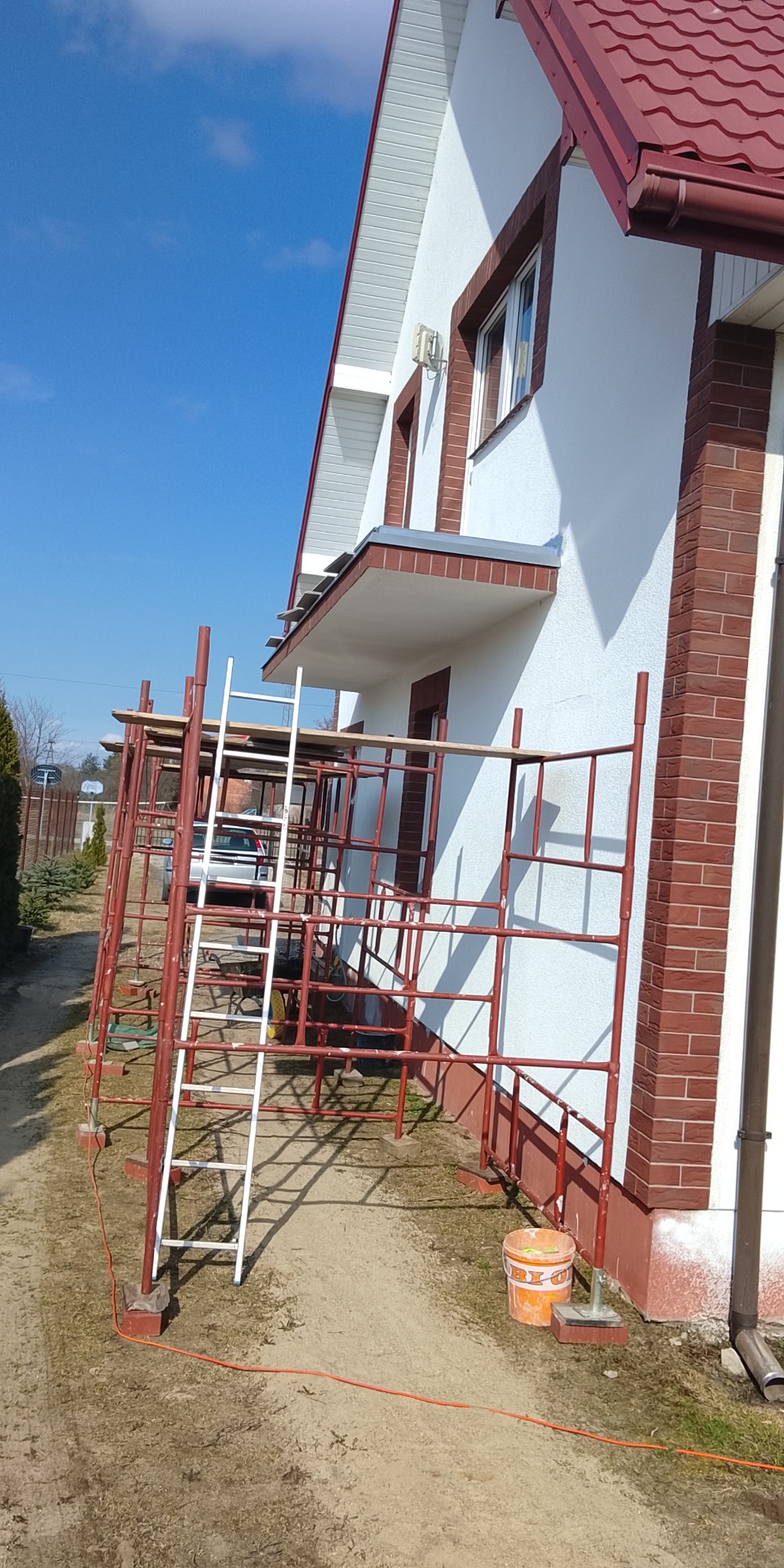 Biały dom z czerwoną cegłą i rusztowaniem. Widoczny fragment elewacji, drabina oparta o rusztowanie, wiadro z farbą. Słoneczny dzień.