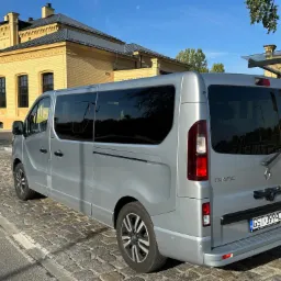 Srebrny Renault Trafic z napisem 'Wynajmij Busa VIP Bus Polska' zaparkowany na brukowanej ulicy przed żółtym budynkiem z cegły, widoczny księżyc na niebie.