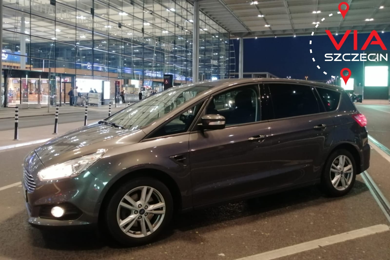 Szary minivan Ford S-Max zaparkowany przed nowoczesnym budynkiem lotniska, z grafiką 'VIA Szczecin' w górnym prawym rogu.