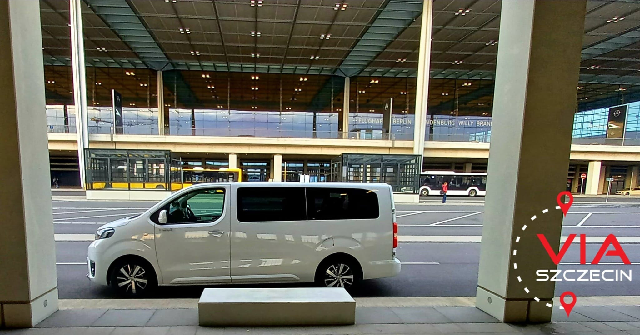 Biały van z przyciemnianymi szybami na tle budynku lotniska Berlin Brandenburg Willy Brandt, widoczny zza kolumn.