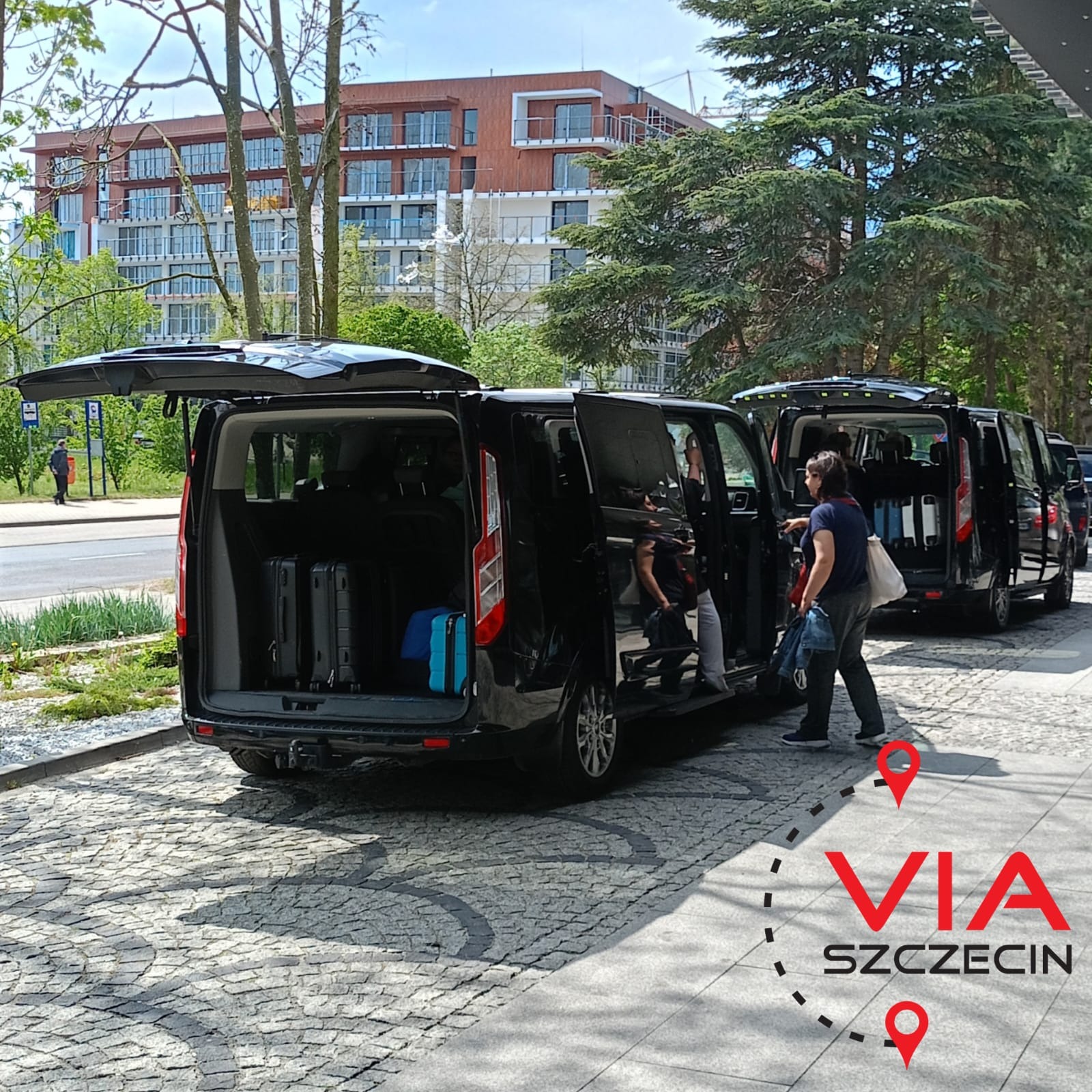 Czarne busy z otwartymi bagażnikami i drzwiami, bagaże w środku, pasażer wsiada, logo VIA Szczecin w prawym dolnym rogu.