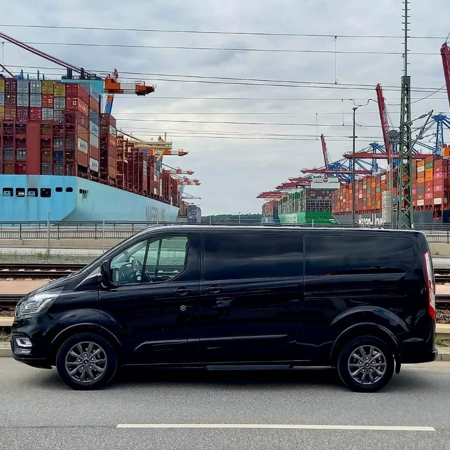 Czarny van na tle portu kontenerowego z suwnicami i statkami, widoczne tory kolejowe.