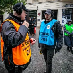 Dwóch stewardów w kamizelkach (pomarańczowej i niebieskiej) z napisem 'Steward' i numerem 204, rozmawia przez krótkofalówki na ulicy w deszczowy dzień. W tle widać przechodniów i budynki.