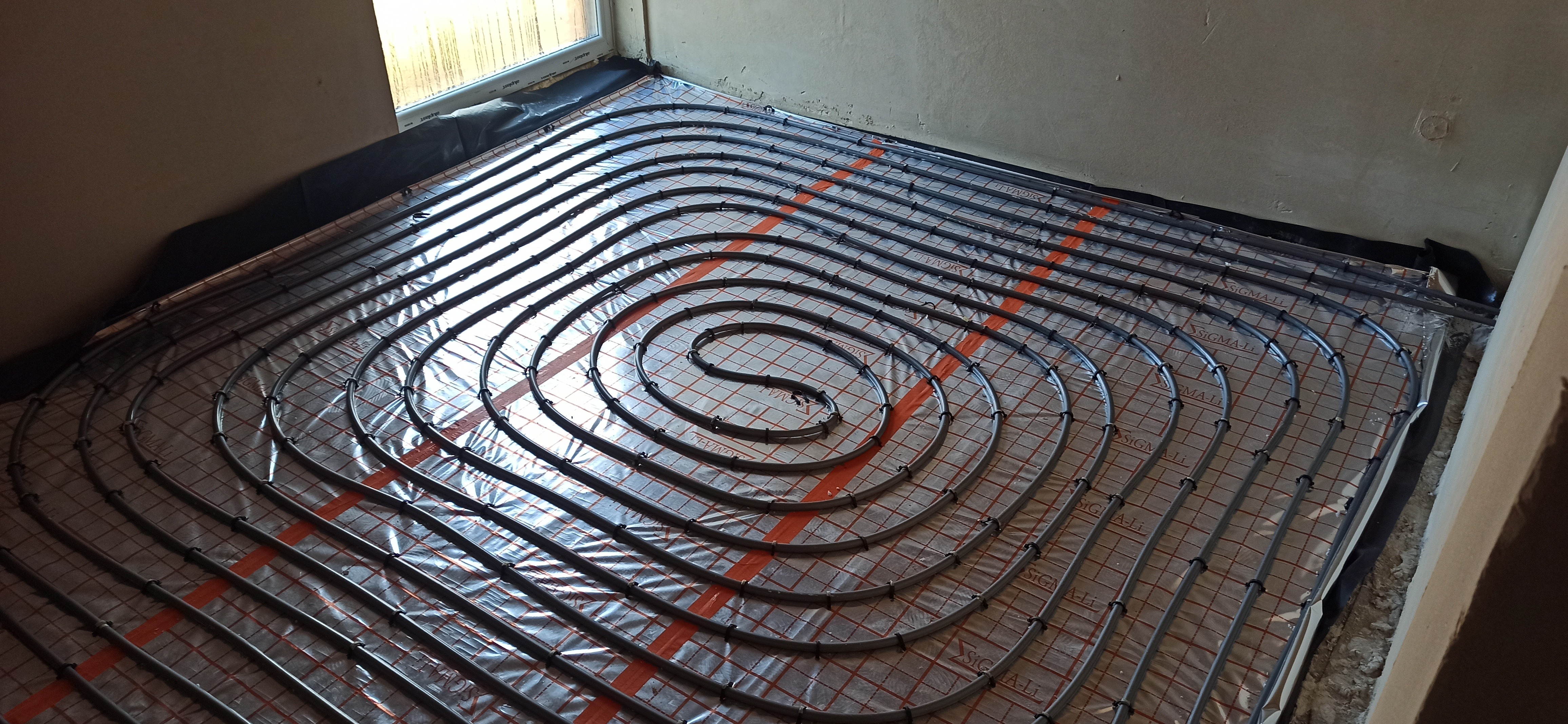 Rozłożona instalacja ogrzewania podłogowego z szarych rurek na folii z czerwoną kratką, widok z góry w surowym wnętrzu.
