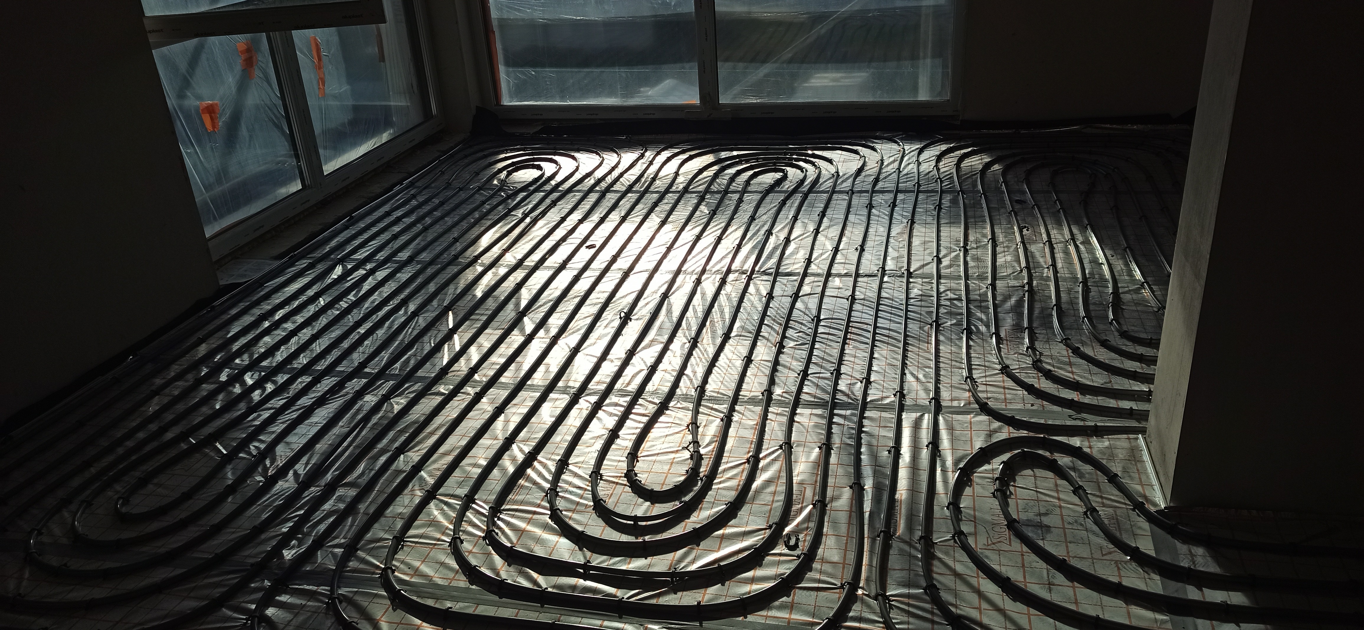 Rozłożone rury ogrzewania podłogowego na folii z siatką, widoczne okno z folią ochronną i fragment ściany w surowym stanie.