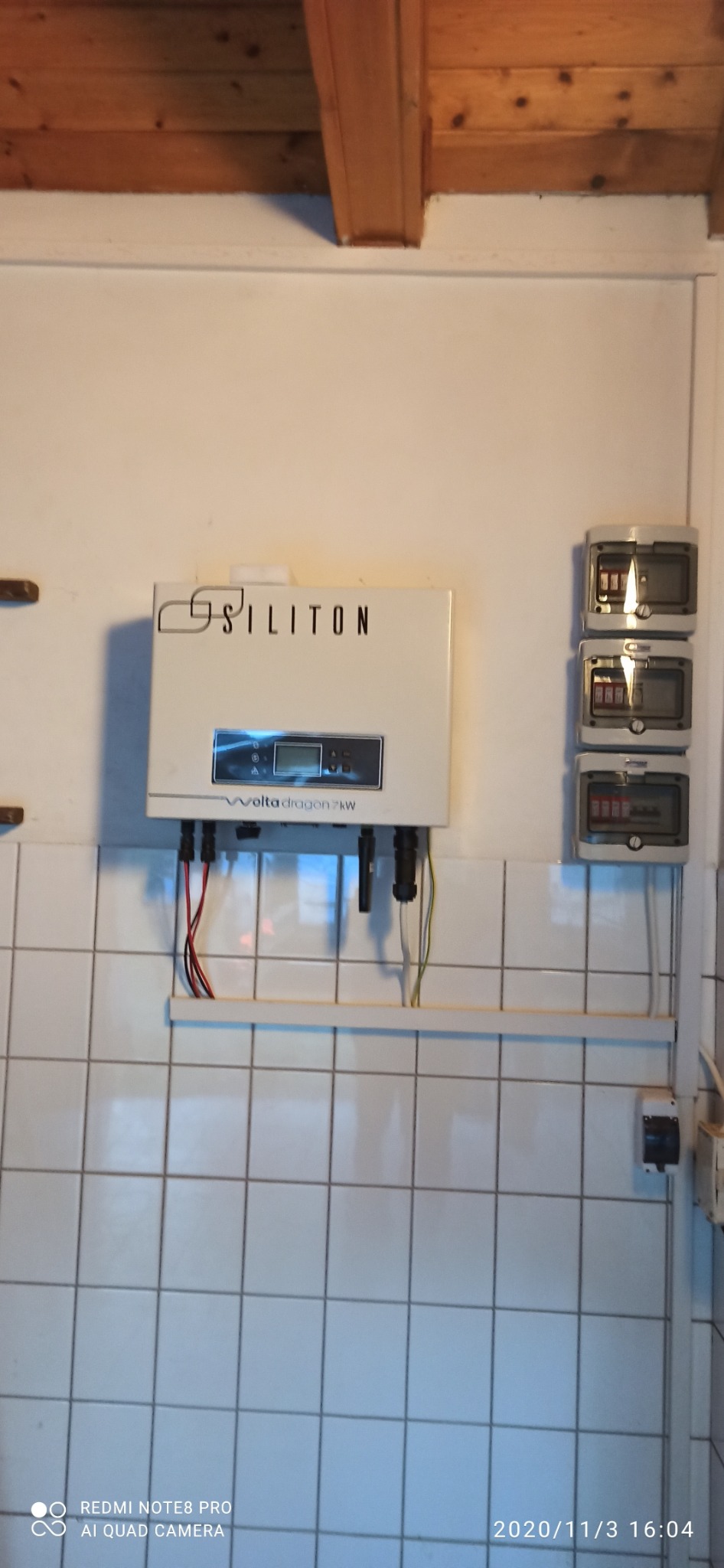 Biały inwerter fotowoltaiczny Siliton Volta Dragon 7kW zamontowany na ścianie z białych płytek, poniżej widoczne kable, obok trzy bezpieczniki.