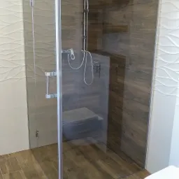 Kabina prysznicowa ze szkła z chromowanymi okuciami, zamontowana w łazience wykończonej płytkami imitującymi drewno i białymi płytkami z falowanym wzorem.