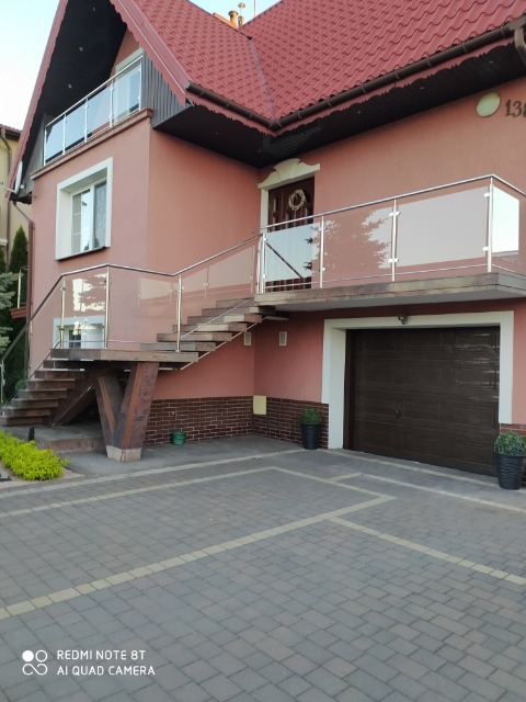 Dom z beżową elewacją, zewnętrznymi schodami z drewnianymi podporami i szklaną balustradą z nierdzewnymi elementami, prowadzącymi na balkon nad garażem.