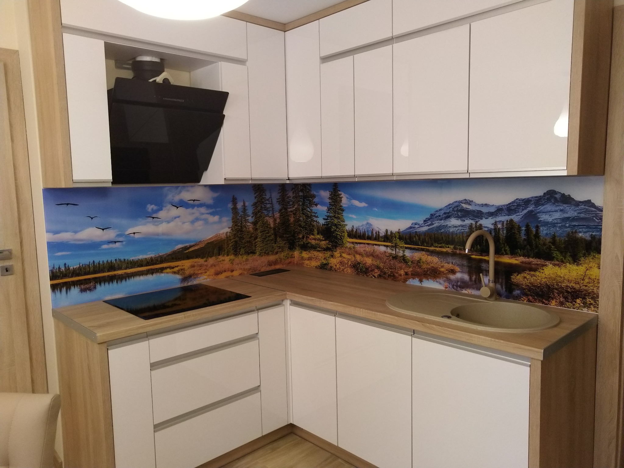 Nowoczesna kuchnia z białymi szafkami i blatem imitującym drewno, ozdobiona panelem szklanym z motywem górskiego krajobrazu z jeziorem i ptakami.