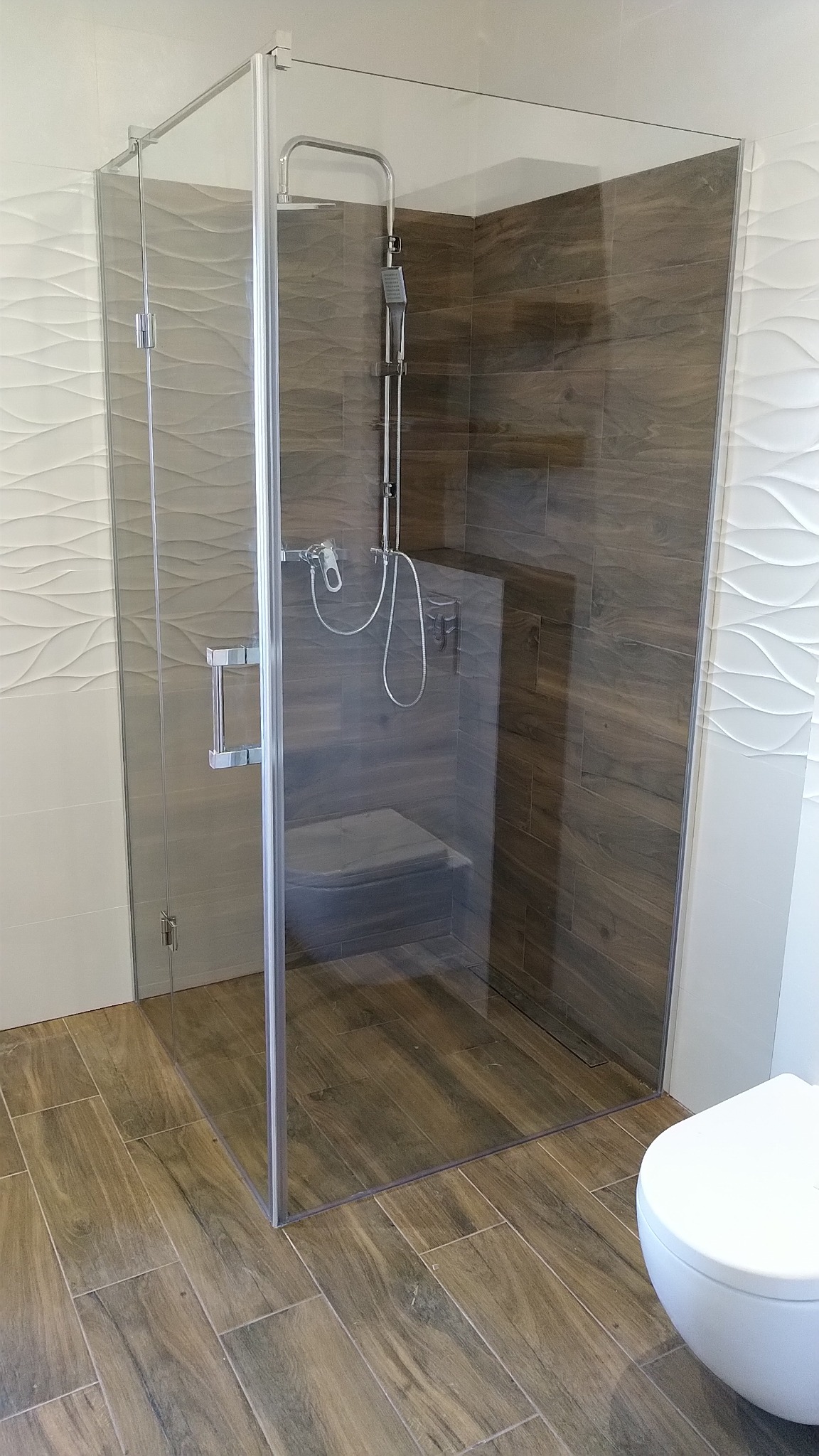 Kabina prysznicowa ze szkła z chromowanymi okuciami, zamontowana w łazience wykończonej płytkami imitującymi drewno i białymi płytkami z falowanym wzorem.