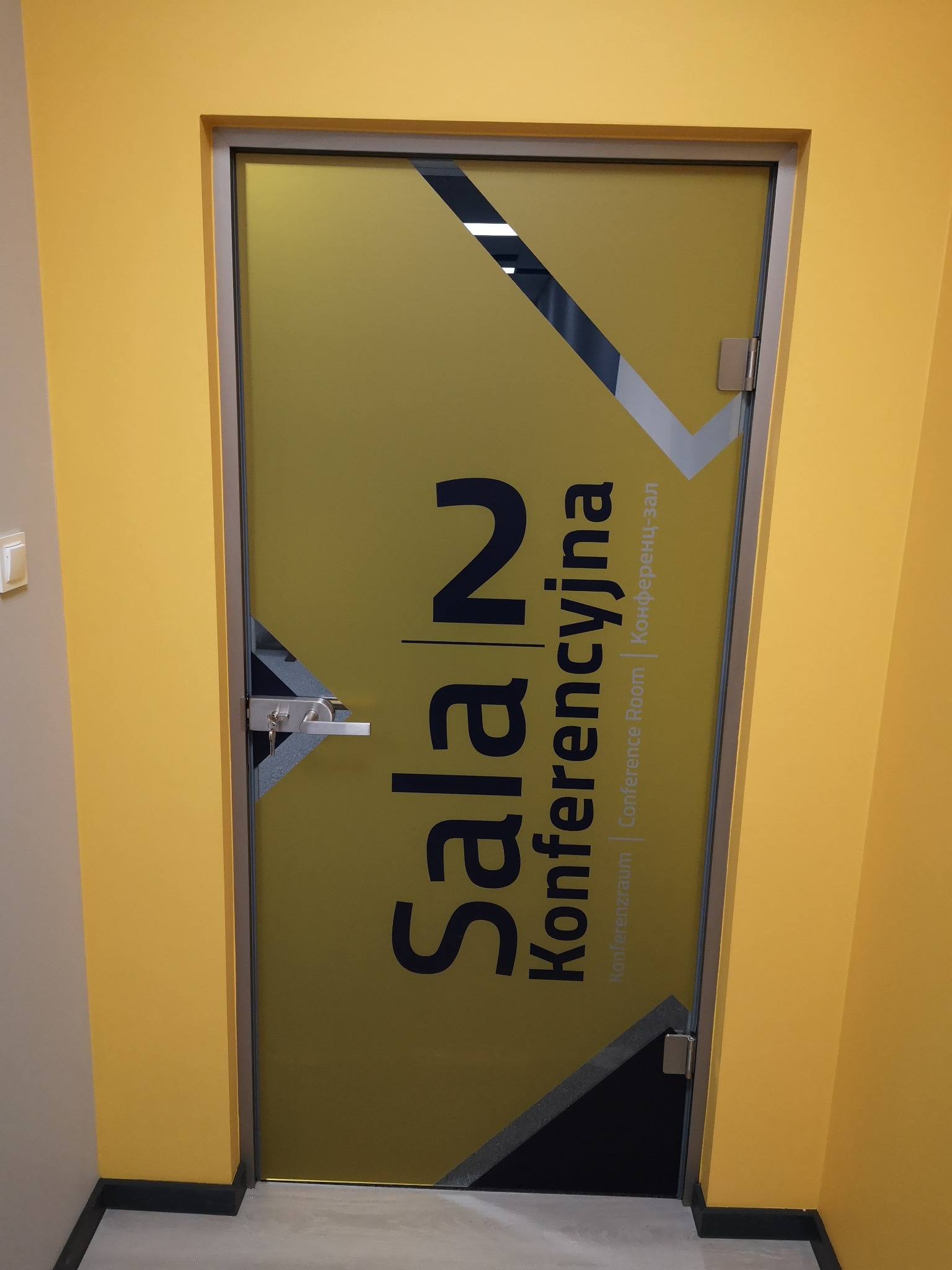 Szklane drzwi do sali konferencyjnej z napisem 'Sala N Konferencyjna' w kolorze czarnym na żółtym tle, widoczny zamek z kluczem, osadzone w żółtej ścianie.