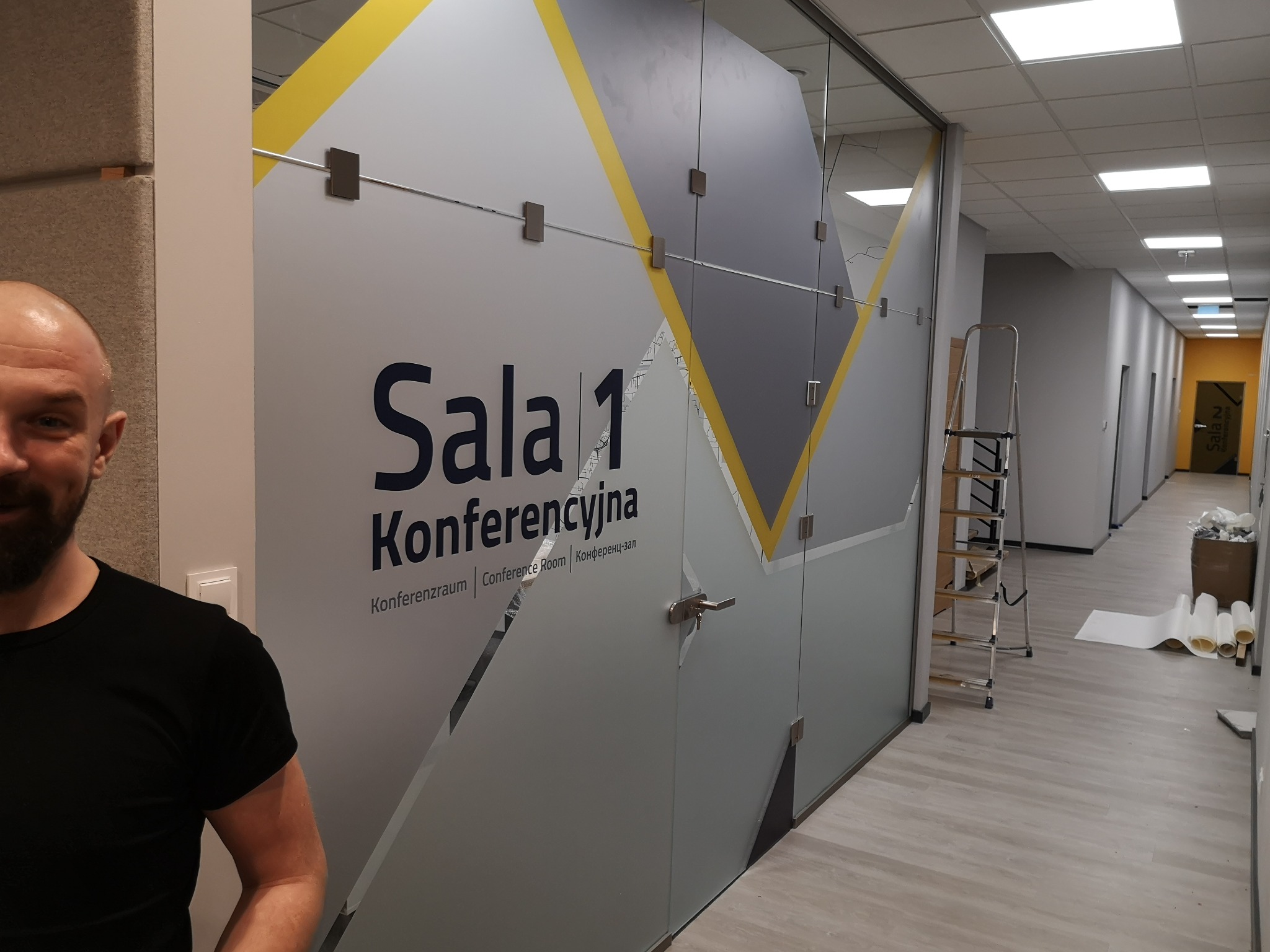 Szklana ściana działowa z dekoracyjną folią z napisem 'Sala 1 Konferencyjna' w biurze, widoczne mocowania i klamka, w tle drabina i materiały montażowe.