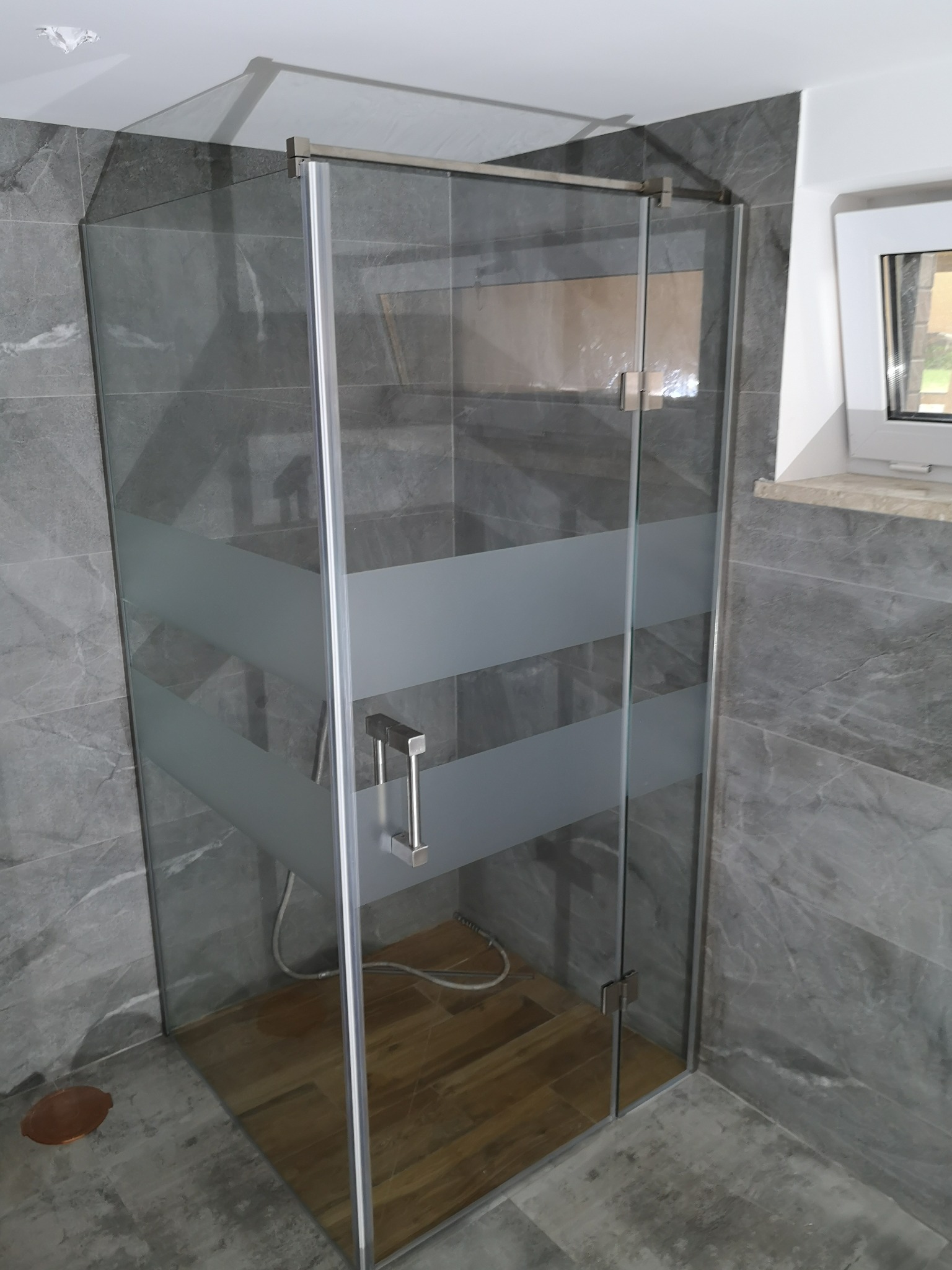 Szklana kabina prysznicowa z matowymi pasami na szybie i chromowanymi okuciami, w łazience wykończonej szarymi płytkami imitującymi marmur, widok z boku.