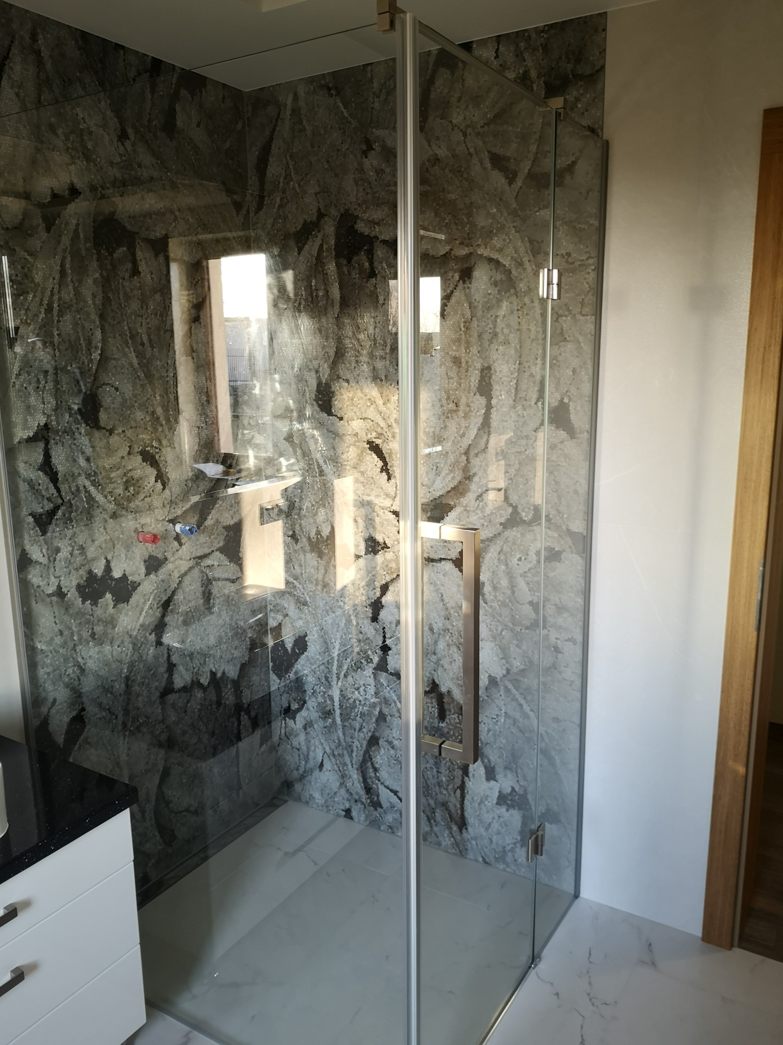 Szklana kabina prysznicowa z chromowanym uchwytem na tle ściany wykończonej płytkami imitującymi marmur.