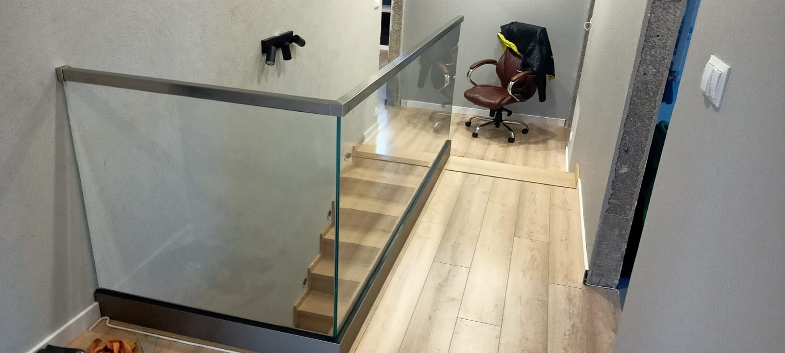 Szklana balustrada z metalową ramą zabezpieczająca drewniane schody wewnętrzne; w tle krzesło biurowe i lampa ścienna.