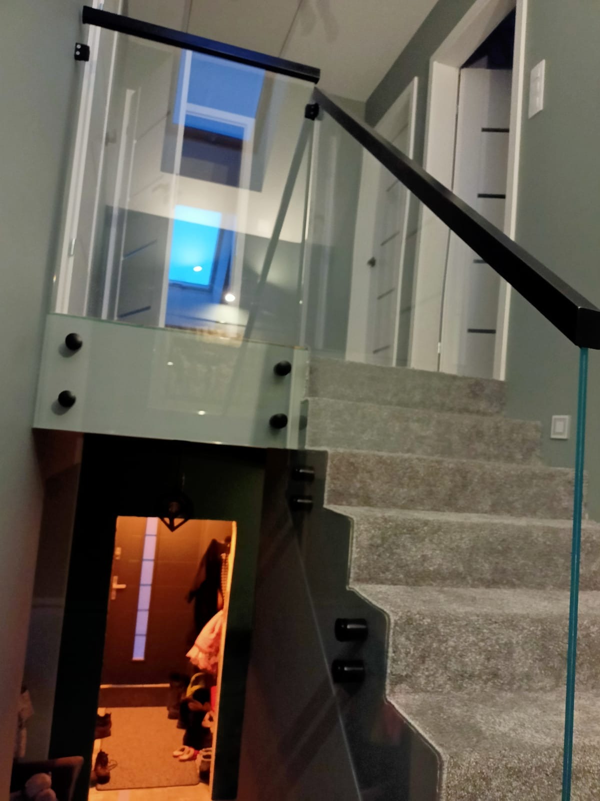 Szklana balustrada z czarną poręczą zamontowana na schodach wewnątrz domu, widok z dołu na oświetlony sufit przez szkło.