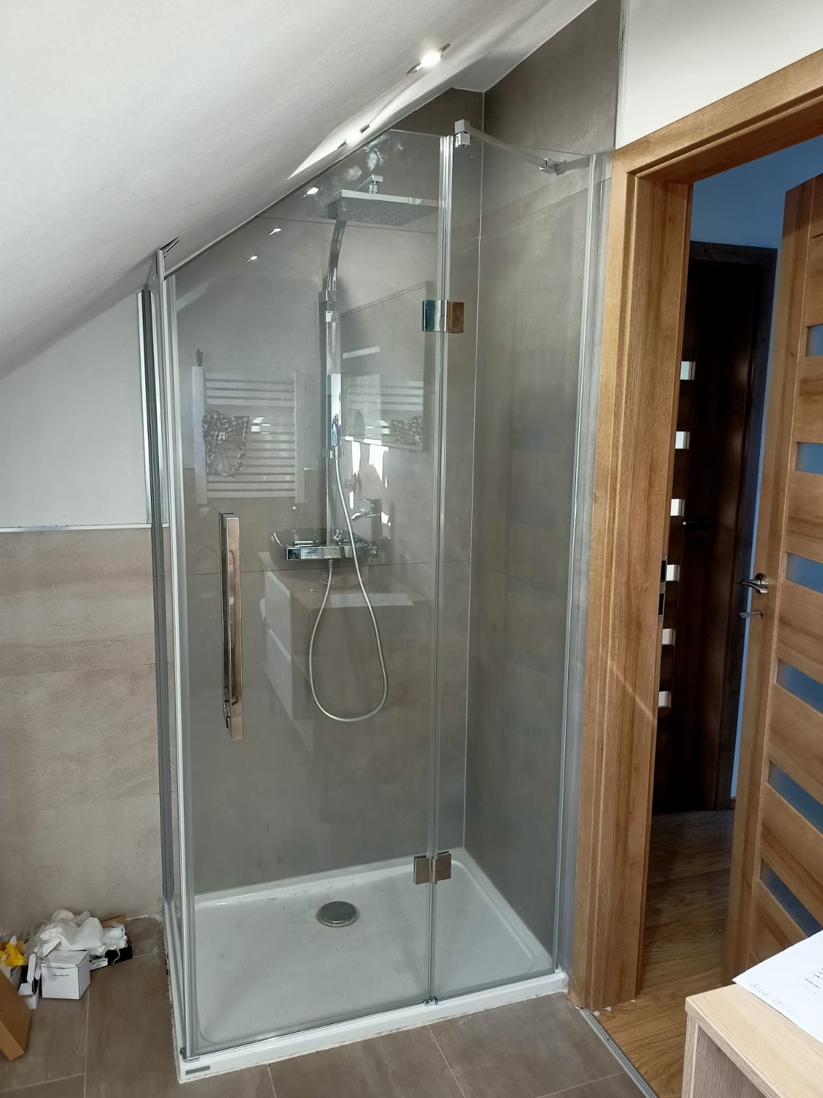 Szklana kabina prysznicowa z chromowanymi zawiasami w łazience na poddaszu, widoczny odpływ w brodziku i fragment drewnianych drzwi.