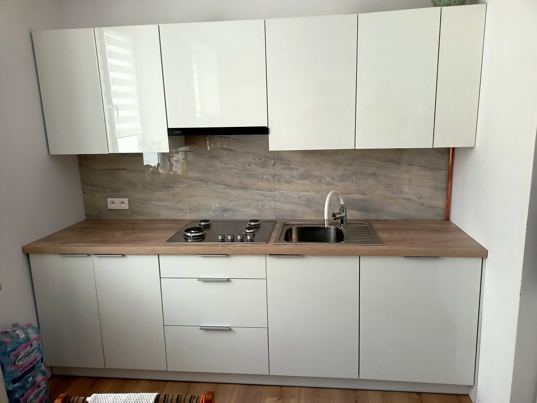 Nowoczesna biała kuchnia z blatem imitującym drewno, z widoczną płytą grzewczą i zlewem ze stali nierdzewnej, marmurowym panelem nad blatem i białymi szafkami wiszącymi.