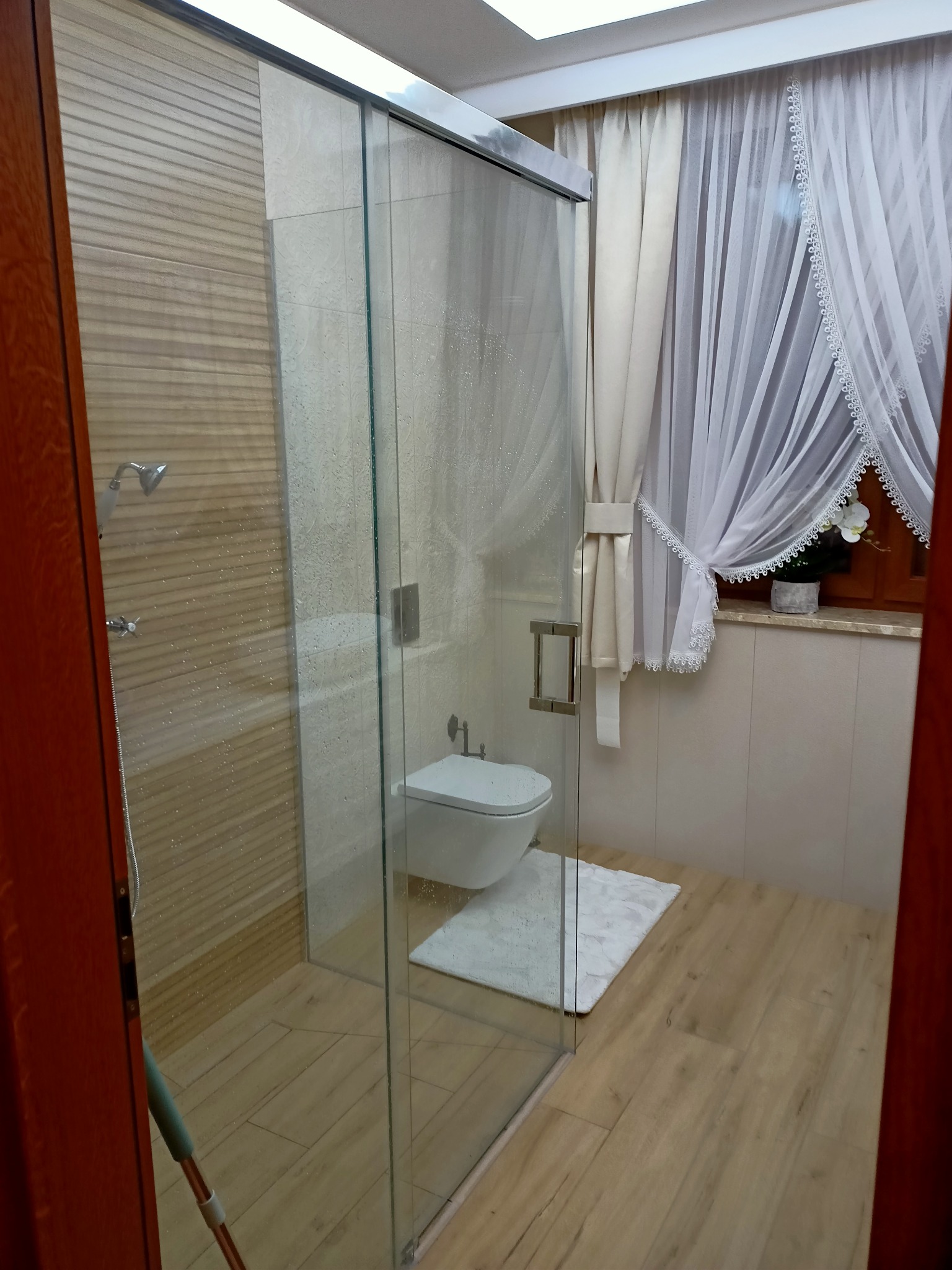 Szklana kabina prysznicowa z przesuwnymi drzwiami, widok z zewnątrz, z widocznym bidetem, białym dywanikiem i oknem z białymi firankami w tle.