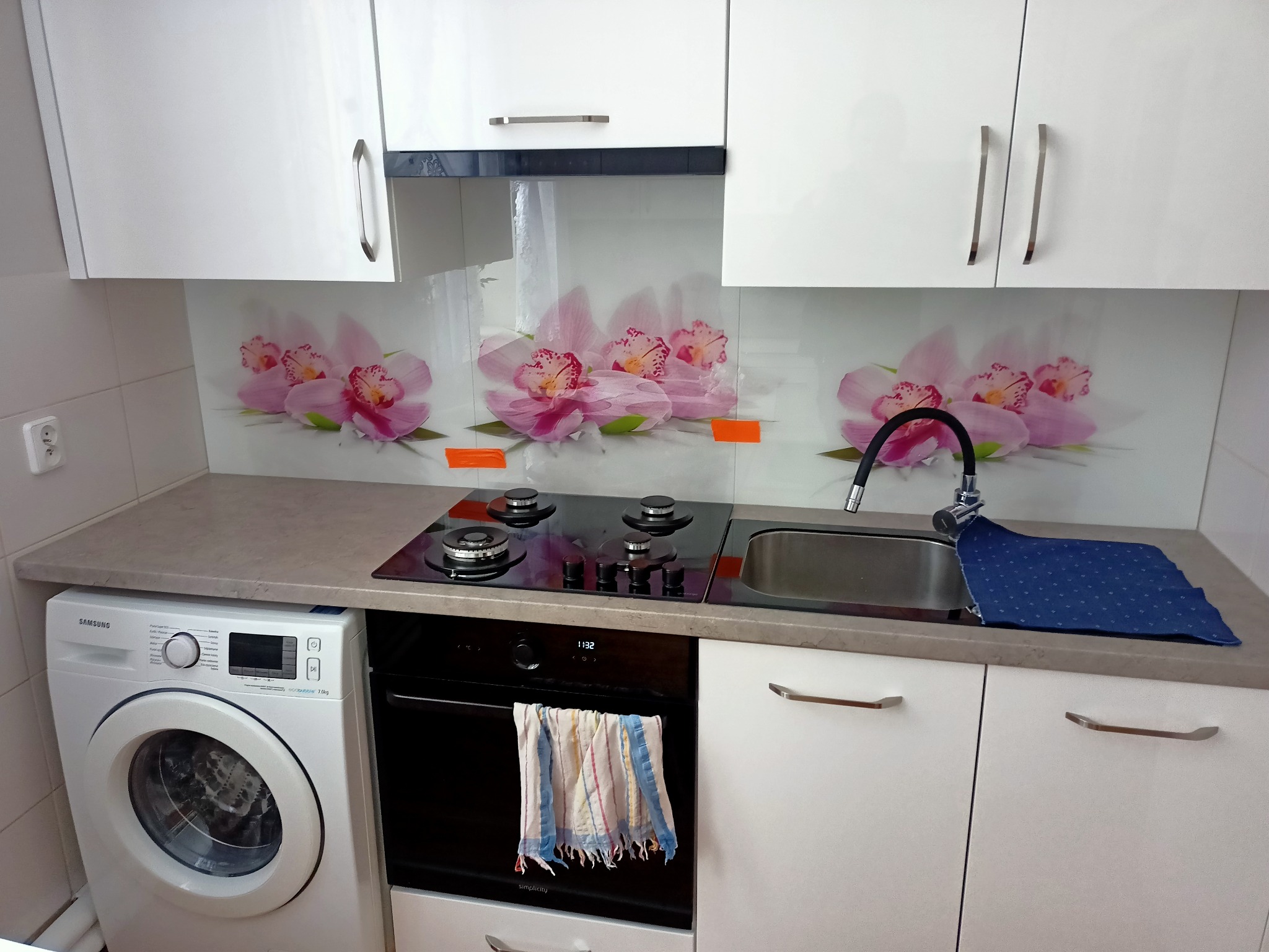 Kuchnia z białymi szafkami, szklanym panelem nad blatem z motywem różowych orchidei, czarną płytą grzewczą i pralką obok. Na piekarniku wisi ściereczka.
