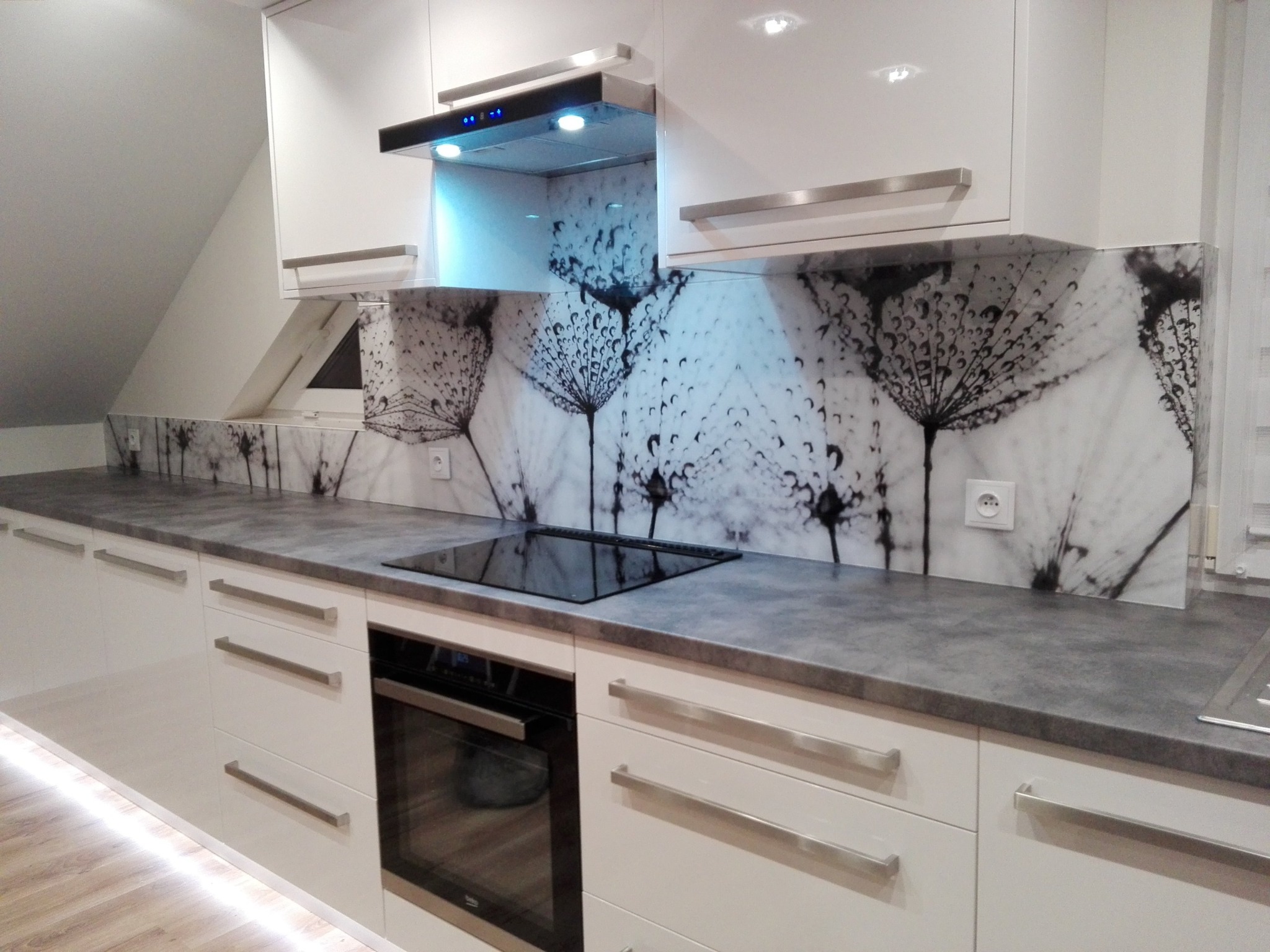 Biała kuchnia z szarym blatem i szklanym panelem ściennym z motywem dmuchawca, oświetlona listwą LED pod szafkami.