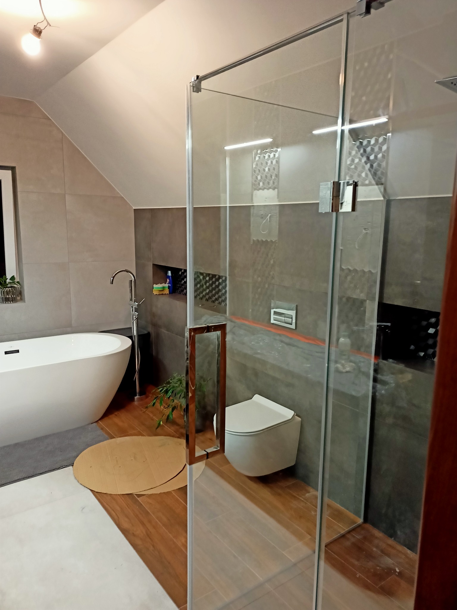 Nowoczesna łazienka na poddaszu z przeszkloną kabiną prysznicową typu walk-in, widoczna biała wanna wolnostojąca, podwieszana toaleta i szare płytki wielkoformatowe.