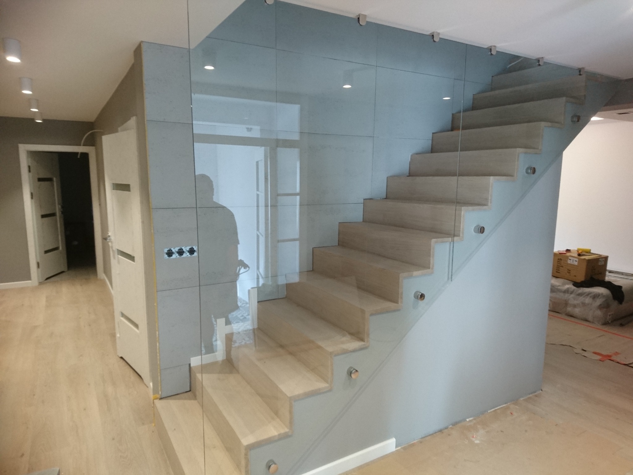 Nowoczesne schody drewniane z przezroczystą balustradą szklaną mocowaną punktowo, widok z dołu na jasne wnętrze z widocznymi drzwiami i podłogą.