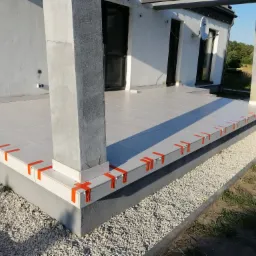 Jasne płytki na tarasie domu jednorodzinnego, zabezpieczone pomarańczową taśmą i klipsami poziomującymi, widok z zewnątrz na fragment elewacji i kolumny podtrzymujące zadaszenie.