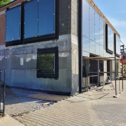 DOOR Filipek sp.k Producent ślusarki i stolarki budowlanej z aluminium i PCV - Montaż okien PCV w surowym budynku, widoczne okna z czarnymi ramami, ślady po tynku i rusztowanie w tle.