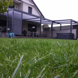 Nowoczesna pergola z regulowanymi lamelami przy domu jednorodzinnym, widok z poziomu trawnika, w tle zestaw mebli ogrodowych i jacuzzi.