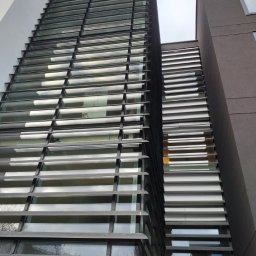 DOOR Filipek sp.k Producent ślusarki i stolarki budowlanej z aluminium i PCV - Wysoki budynek z fasadą z poziomych, metalowych żaluzji zamontowanych na oknach, widok z dołu w pochmurny dzień.