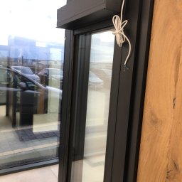 DOOR Filipek sp.k Producent ślusarki i stolarki budowlanej z aluminium i PCV - Ciemna rama okna PCV z roletą zewnętrzną w trakcie montażu, widoczne kable elektryczne, na zewnątrz zaparkowane samochody.