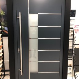 DOOR Filipek sp.k Producent ślusarki i stolarki budowlanej z aluminium i PCV - Nowoczesne, antracytowe drzwi wejściowe z długim, pionowym uchwytem i przeszklonym panelem z poziomymi, srebrnymi akcentami, prezentowane w salonie sprzedaży.