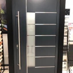 Nowoczesne, antracytowe drzwi wejściowe z długim, pionowym uchwytem i przeszklonym panelem z poziomymi, srebrnymi akcentami, prezentowane w salonie sprzedaży.