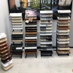 DOOR Filipek sp.k Producent ślusarki i stolarki budowlanej z aluminium i PCV - Ekspozycja różnorodnych próbek parapetów: drewnianych, z konglomeratu i innych materiałów, ułożonych na stojakach w salonie wystawowym, z plakatem reklamowym w tle.