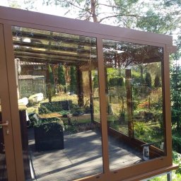 DOOR Filipek sp.k Producent ślusarki i stolarki budowlanej z aluminium i PCV - Zabudowa tarasu z brązowymi ramami aluminiowymi i szklanymi panelami, widok na ogród z drewnianym tarasem i meblami ogrodowymi.