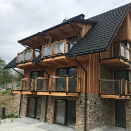 Nowoczesny dom z drewnianą elewacją i kamienną podmurówką, balkony z szklanymi balustradami, czarne rynny i okna, widok z dołu na tle pochmurnego nieba.