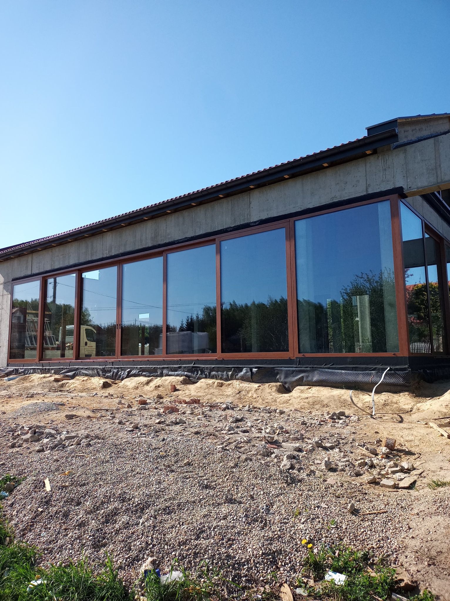 Nowoczesna fasada budynku z dużymi przesuwnymi oknami w brązowych ramach, montowanymi w Bochni, widoczna na tle nieba i częściowo zagospodarowanego terenu.