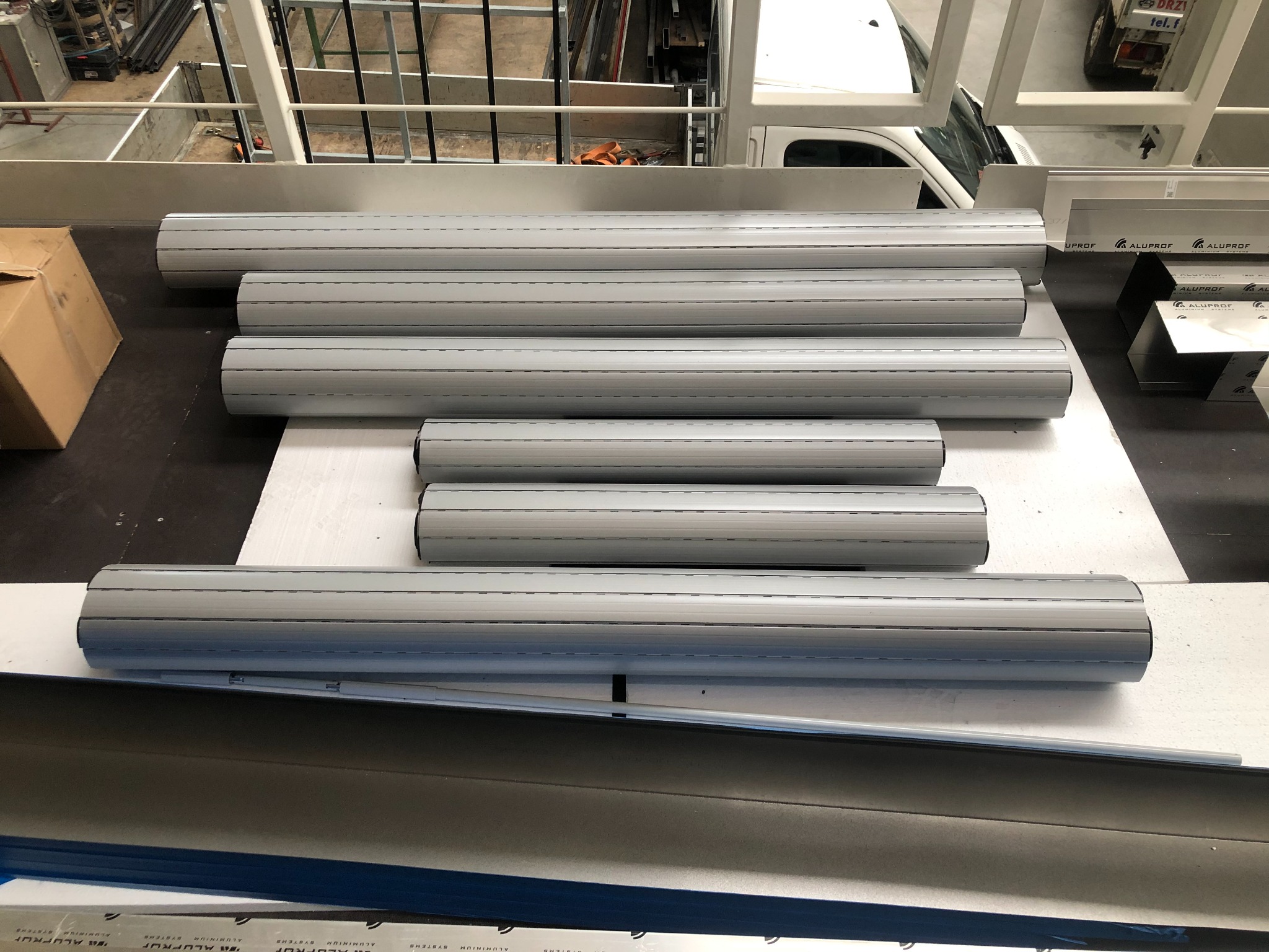 Przygotowane do montażu aluminiowe profile rolet zewnętrznych w warsztacie, widoczny karton, elementy konstrukcji stalowej i częściowo biały samochód dostawczy w tle.