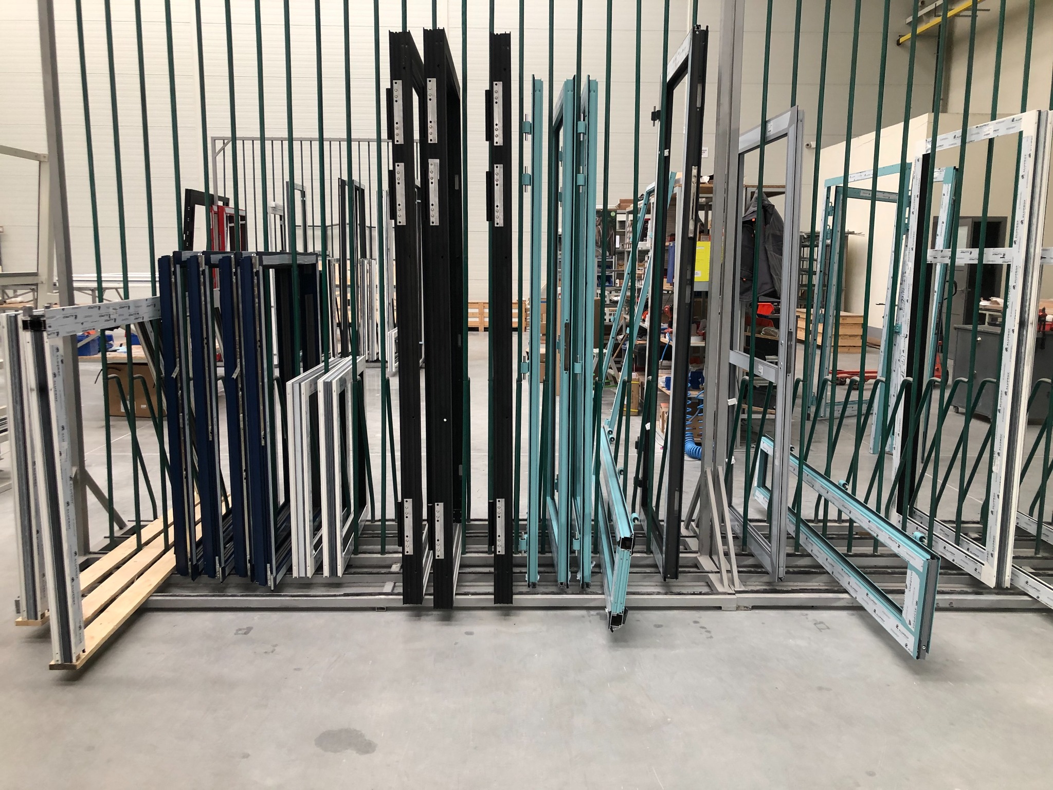 Różnokolorowe ramy okienne, niektóre z okuciami, ustawione pionowo w metalowym stojaku w hali produkcyjnej.