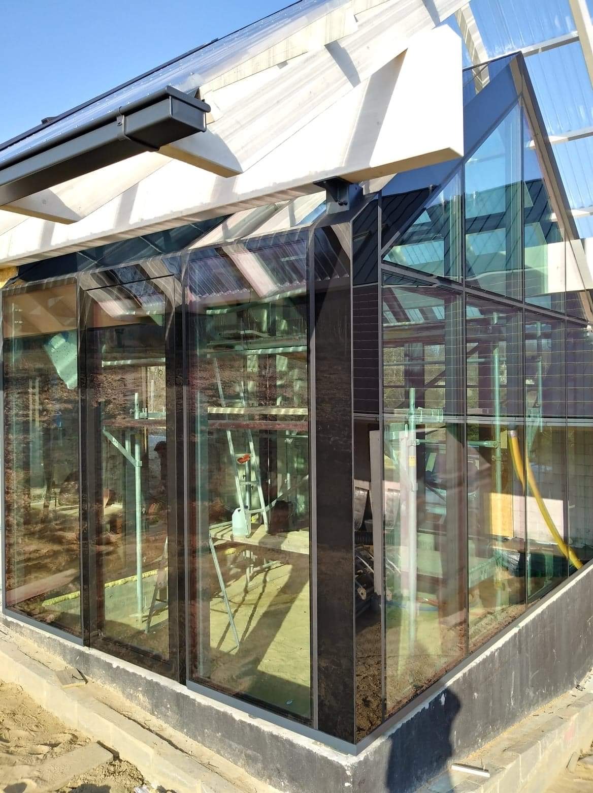 Szklana konstrukcja ogrodu zimowego z czarnymi profilami i drewnianym szkieletem dachu, widoczne rusztowanie wewnątrz, w trakcie montażu.