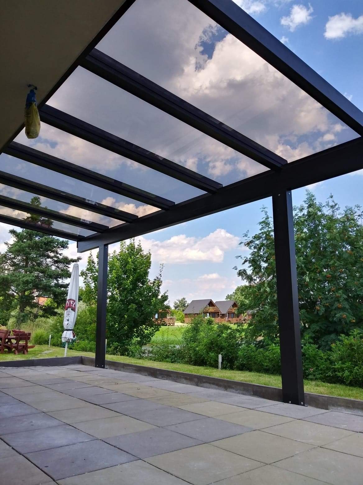 Zadaszenie tarasu z czarną konstrukcją i przezroczystym dachem, odbijającym błękitne niebo z chmurami, widok na ogród z domkami letniskowymi w tle.