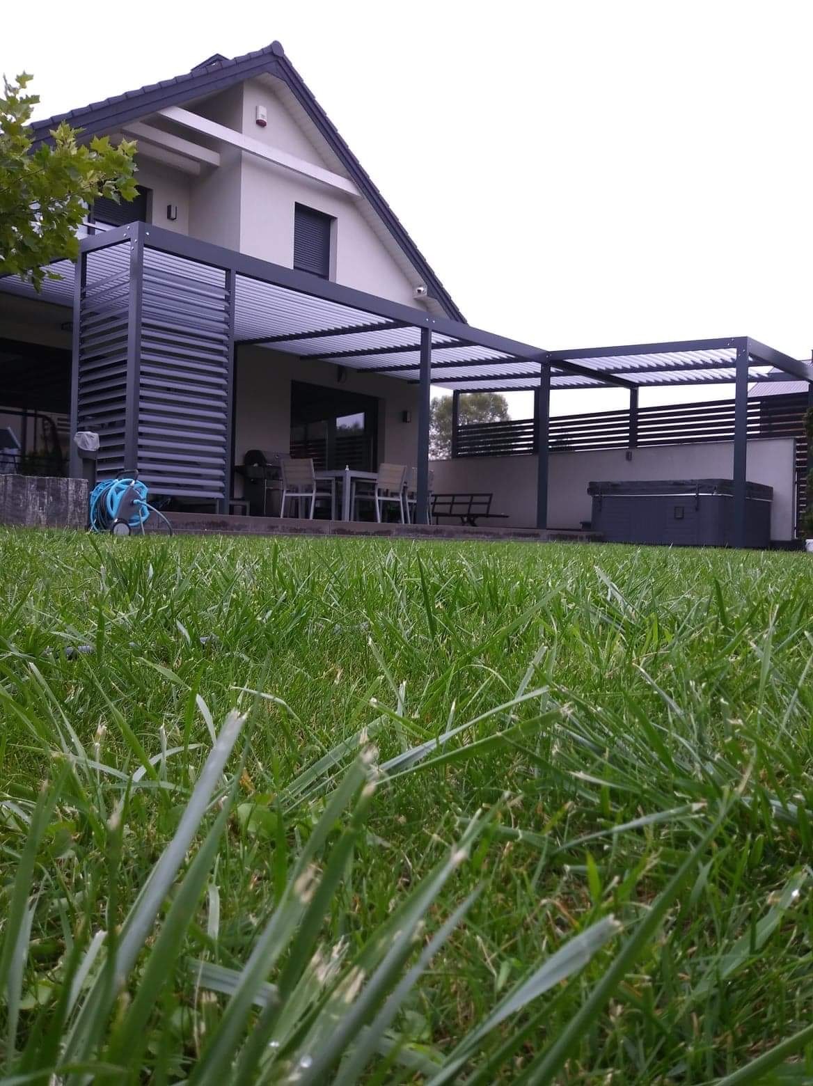 Nowoczesna pergola z regulowanymi lamelami przy domu jednorodzinnym, widok z poziomu trawnika, w tle zestaw mebli ogrodowych i jacuzzi.